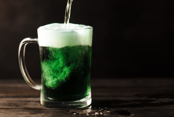 alt = "Clear glass mug filled with green colored beer for Celebrating St. Patrick's Day in NY".