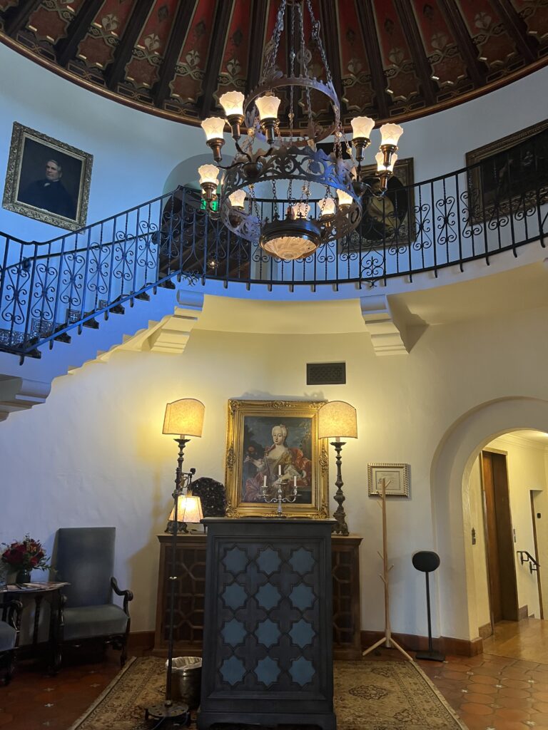 alt txt = "Grand winding staircase with Black handrails and a light fixture inside the Wrigley mansion." 