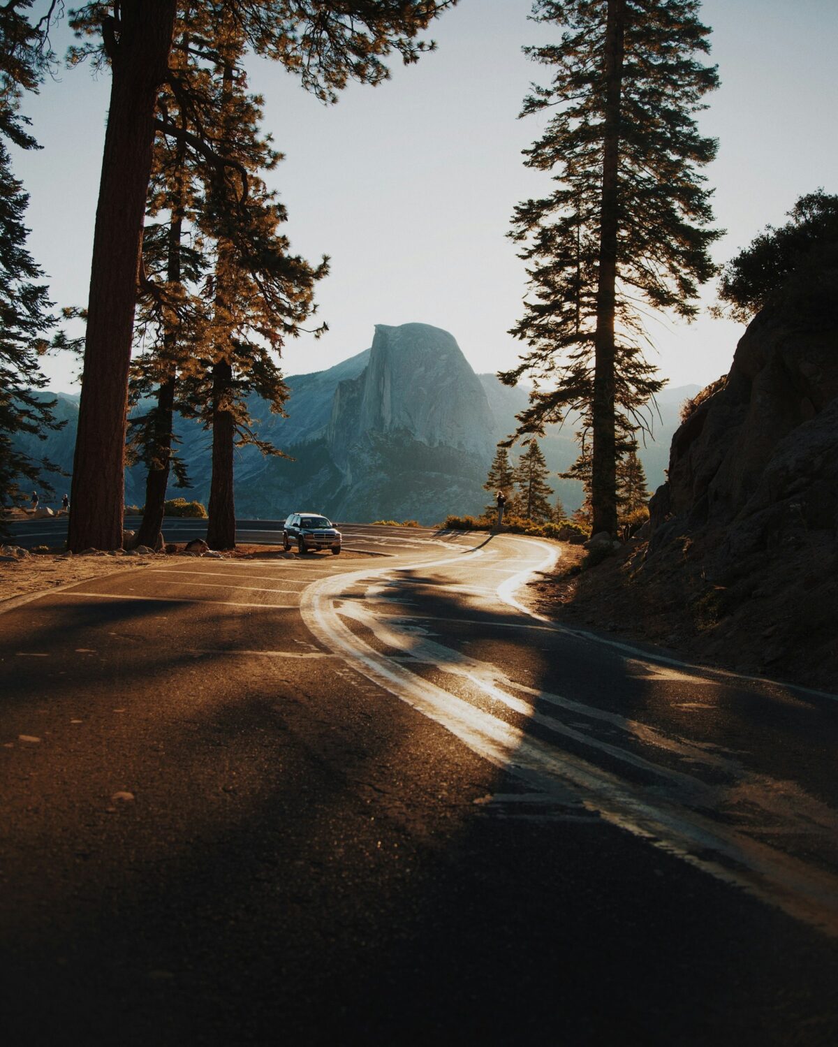 alt txt = "Lone car driving on a winding road surrounded by the sunrise and tall trees in California."