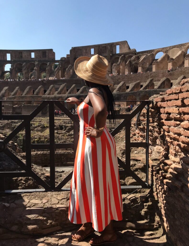 alt txt = "African American woman wearing an orange and white striped dress surrounded by the Colosseum in Italy."