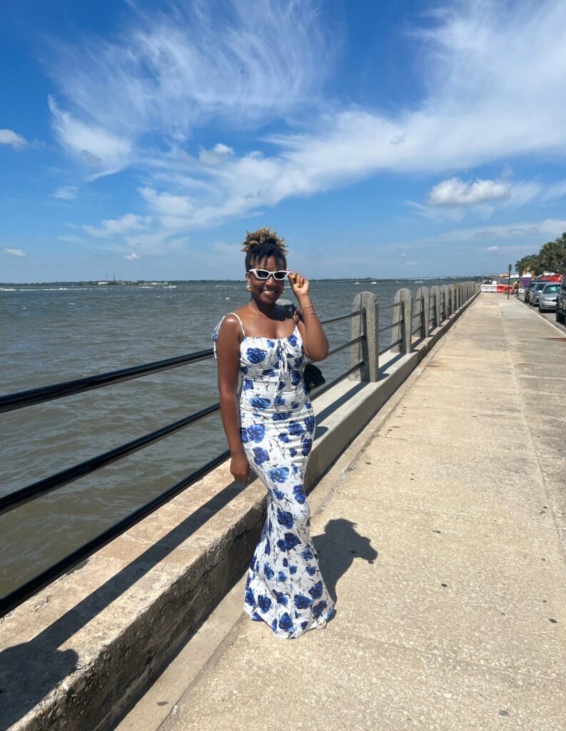 alt txt = "African American woman standing by the ocean wearing a white and blue dress with flowers."