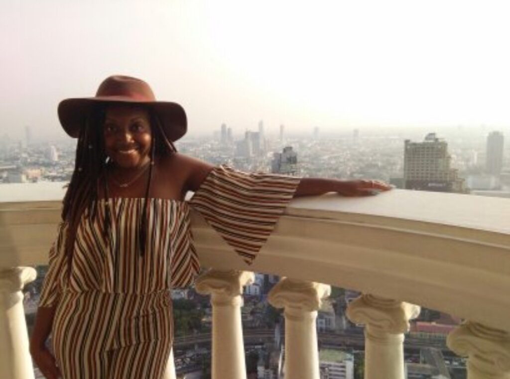 alt txt = "African American woman wearing a striped brown jumpsuit and brown hat standing on a balcony."