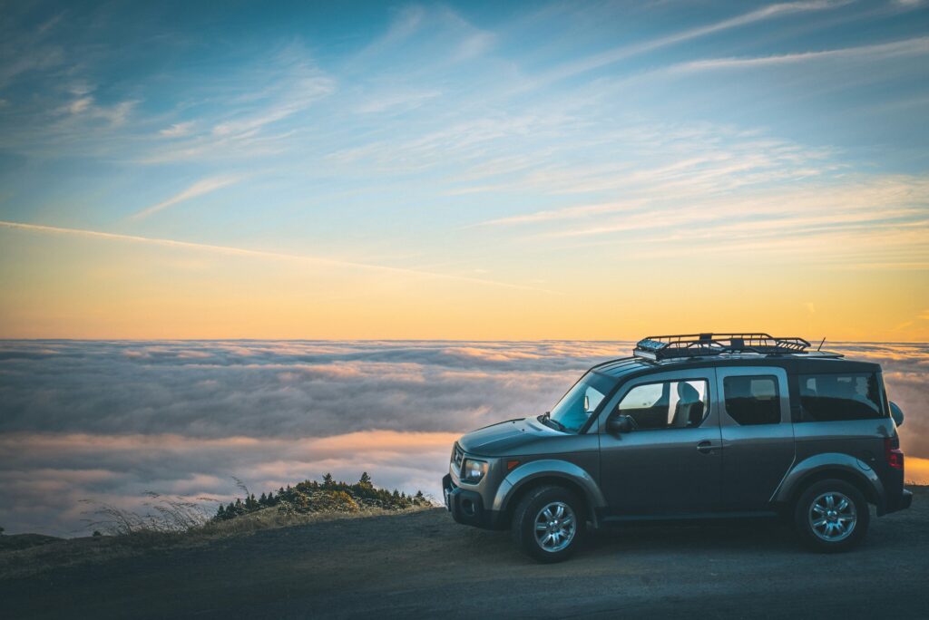 alt txt = "Grey jeep parked on an empty road surrounded by blue skies and clouds".