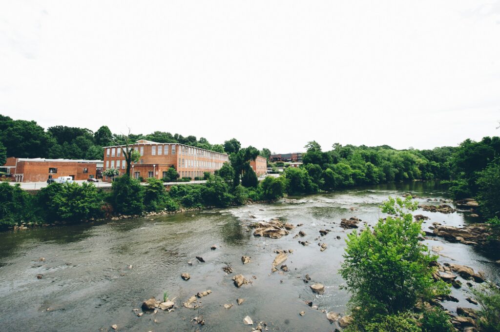 alt txt = "Red brick building surrounded by greenery and a lake with rocks in Saxapahaw, North Carolina".