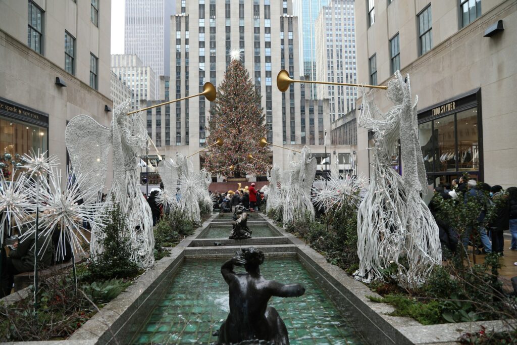 alt txt = "Decorated Christmas tree surrounded by tall buildings, black statues and a water pond in New York City".