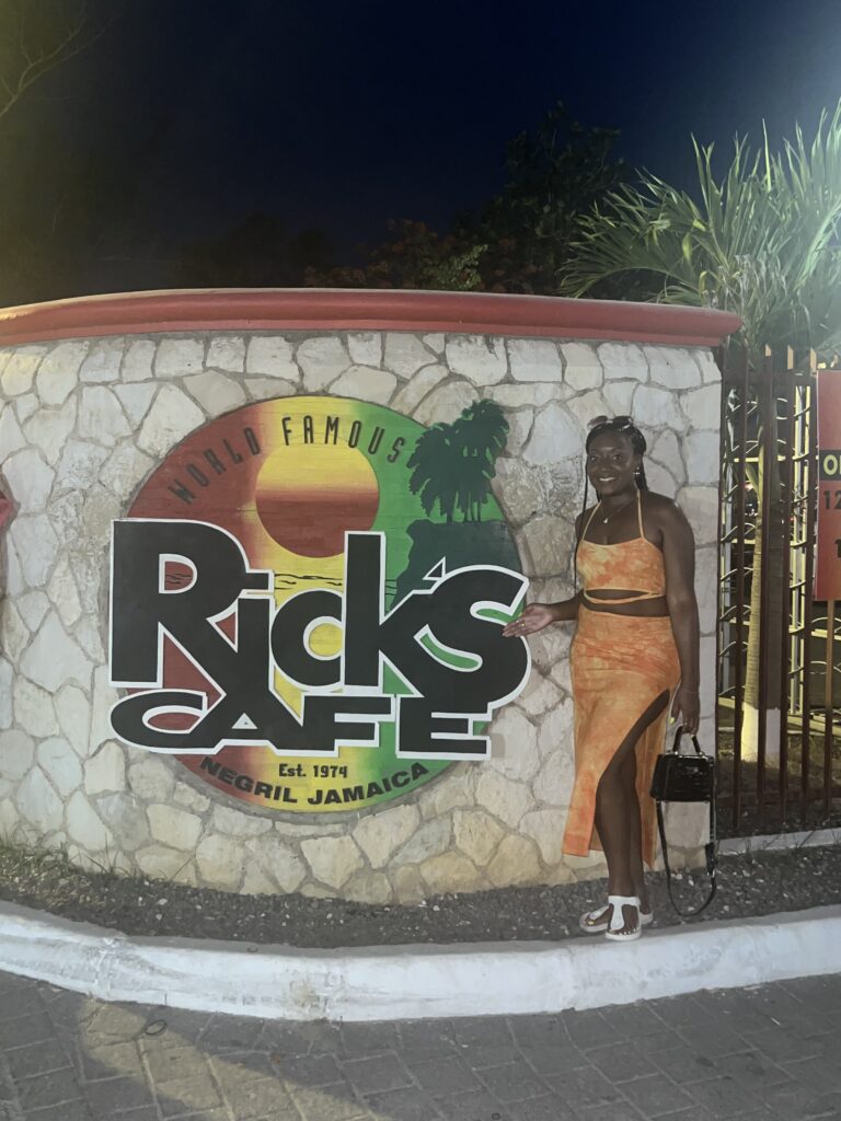 alt txt = " African American woman wearing a two piece orange suit facing a large green, red and yellow restaurant sign".