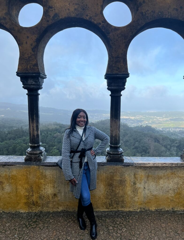 alt txt = "African American woman wearing a black and white coat standing in front of a multicolored castle".