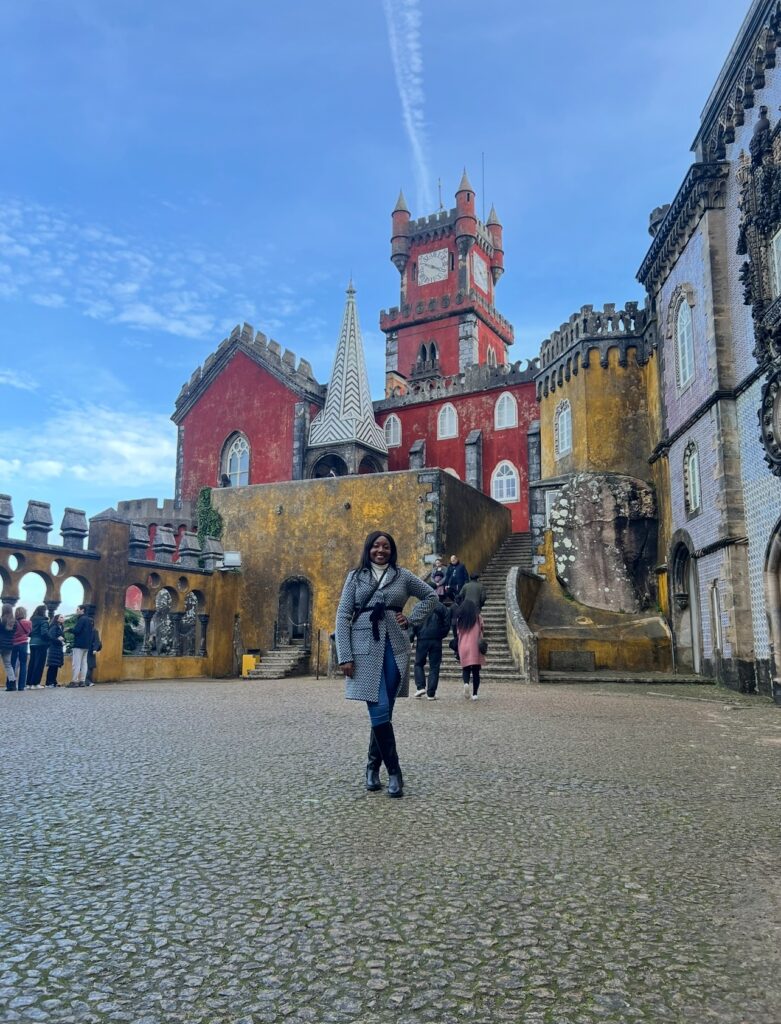 alt txt = "African American woman wearing a black and white coat standing in front of a multicolored castle".