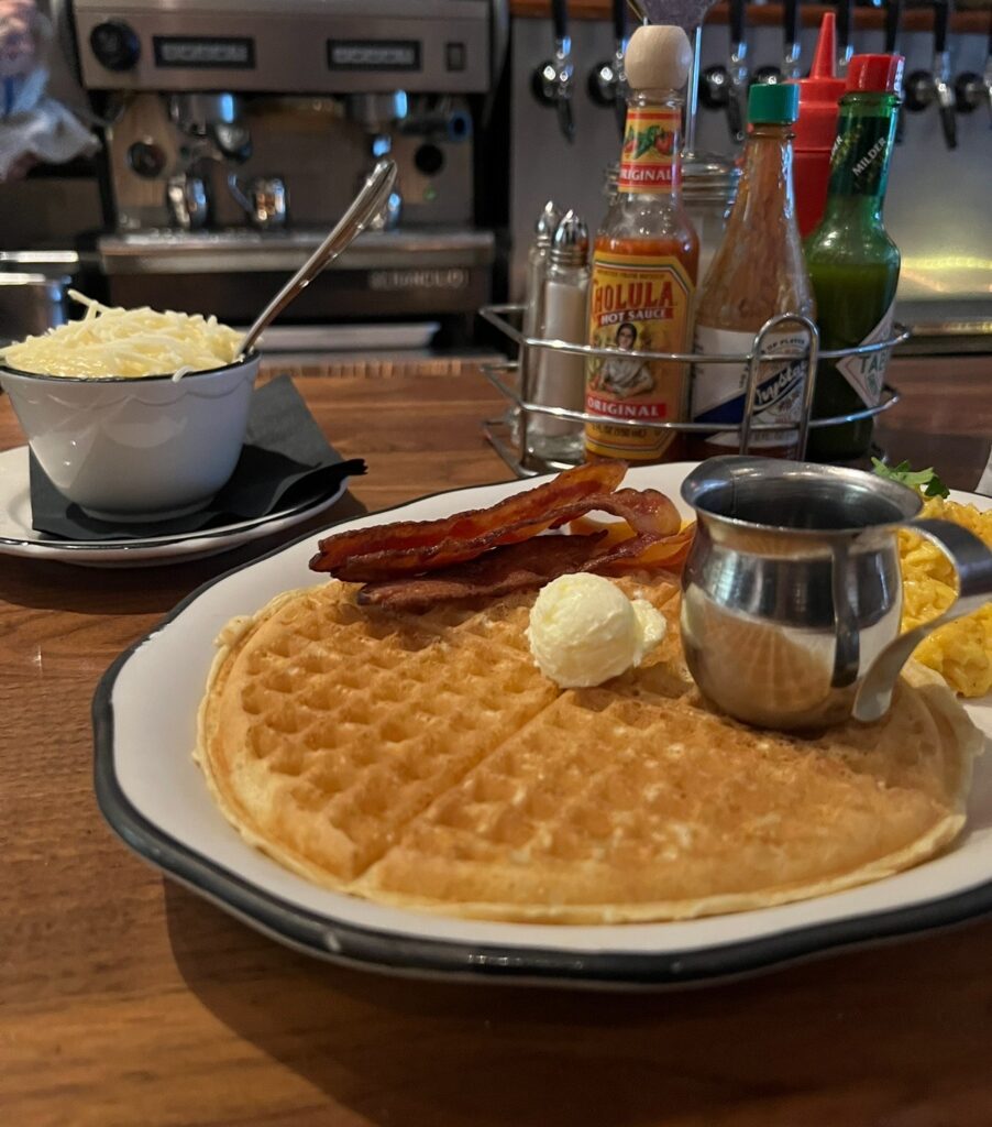 A breakfast plate with a large waffle topped with butter and syrup, crispy bacon, scrambled eggs, and a bowl of cheese grits on the side, set on a wooden counter with hot sauces in the background.