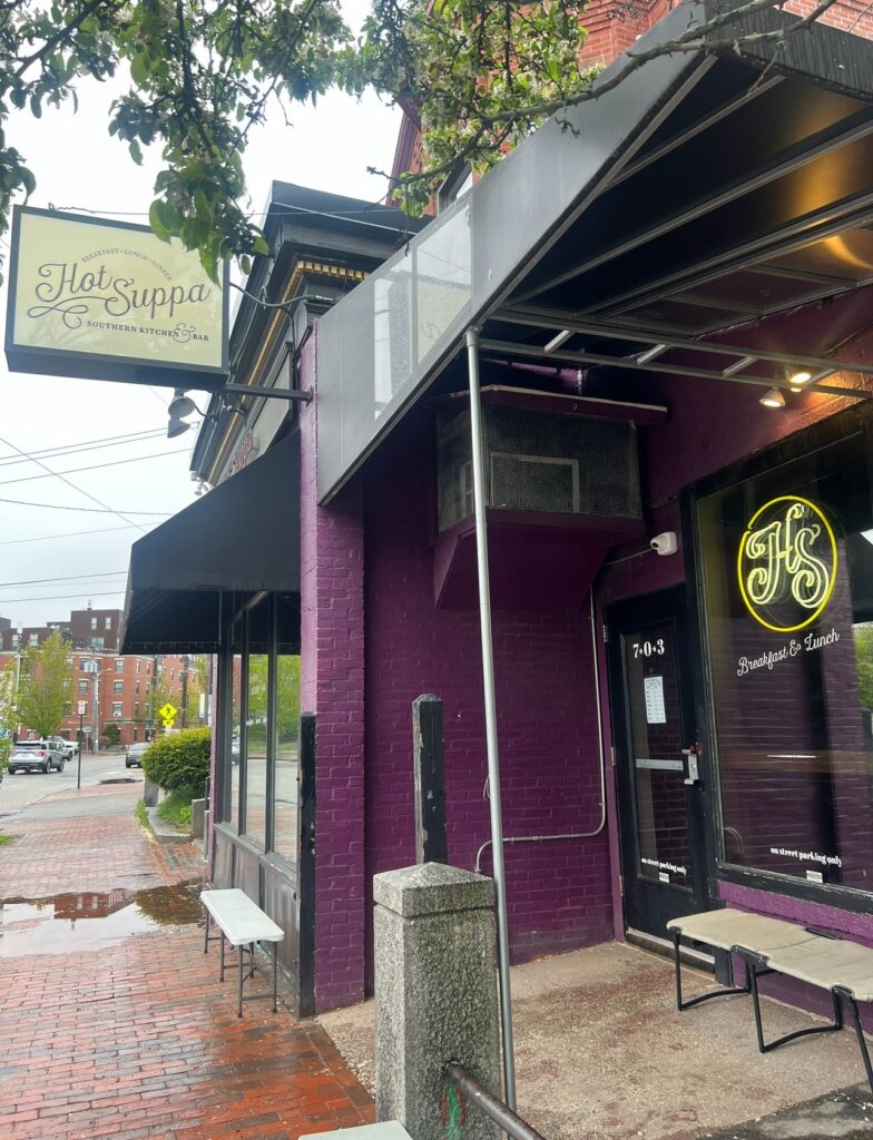 Exterior view of Hot Suppa restaurant in Portland, Maine, featuring a deep purple brick façade, black awnings, and a vintage-style hanging sign under a tree-lined street.