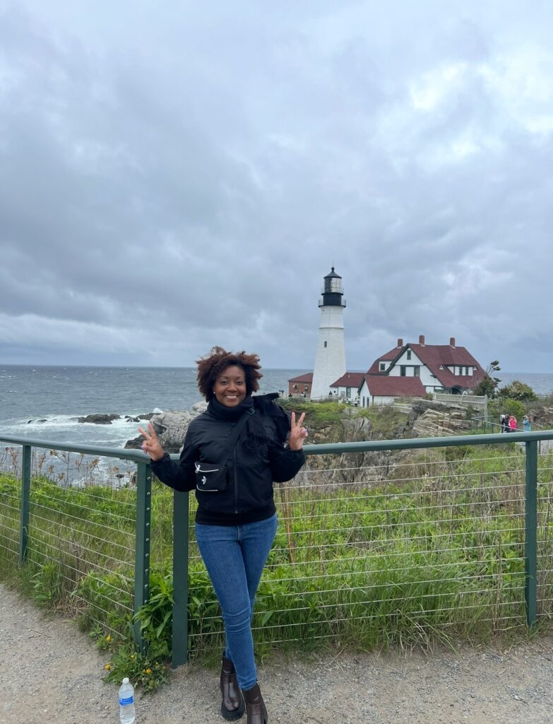 A smiling African American woman in a black jacket and jeans poses with a peace sign in front of a scenic overlook of Portland Head Light. The ocean and lighthouse are visible in the background, with wild grass and a wire fence in the foreground.