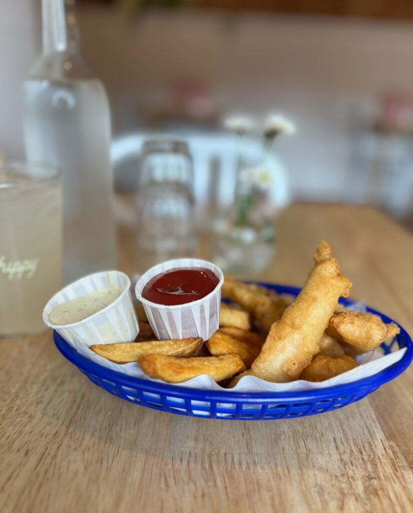 A close-up of a blue plastic basket filled with golden, crispy fish and thick-cut fries served with ketchup and tartar sauce in paper cups. The meal is placed on a wooden table with a water bottle, lemonade, and glassware in the softly blurred background.