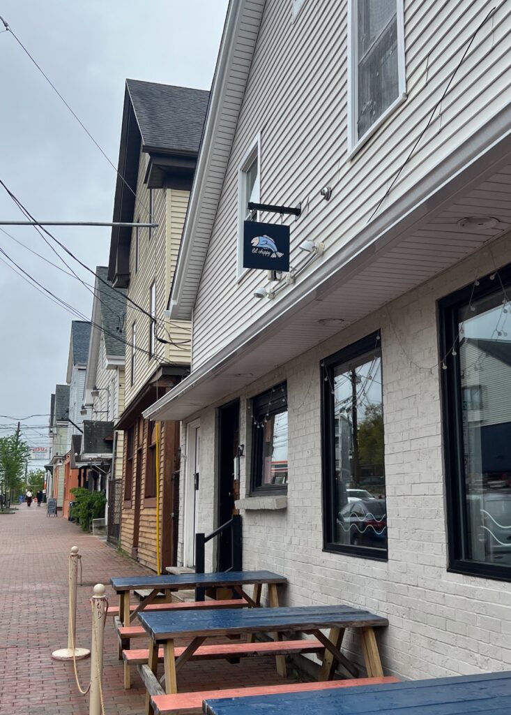 A street view of the Lil' Chippy restaurant, identified by a small hanging sign featuring a stylized fish logo. The white-painted building has black-framed windows and picnic-style benches outside along a quiet brick sidewalk lined with neighboring buildings.