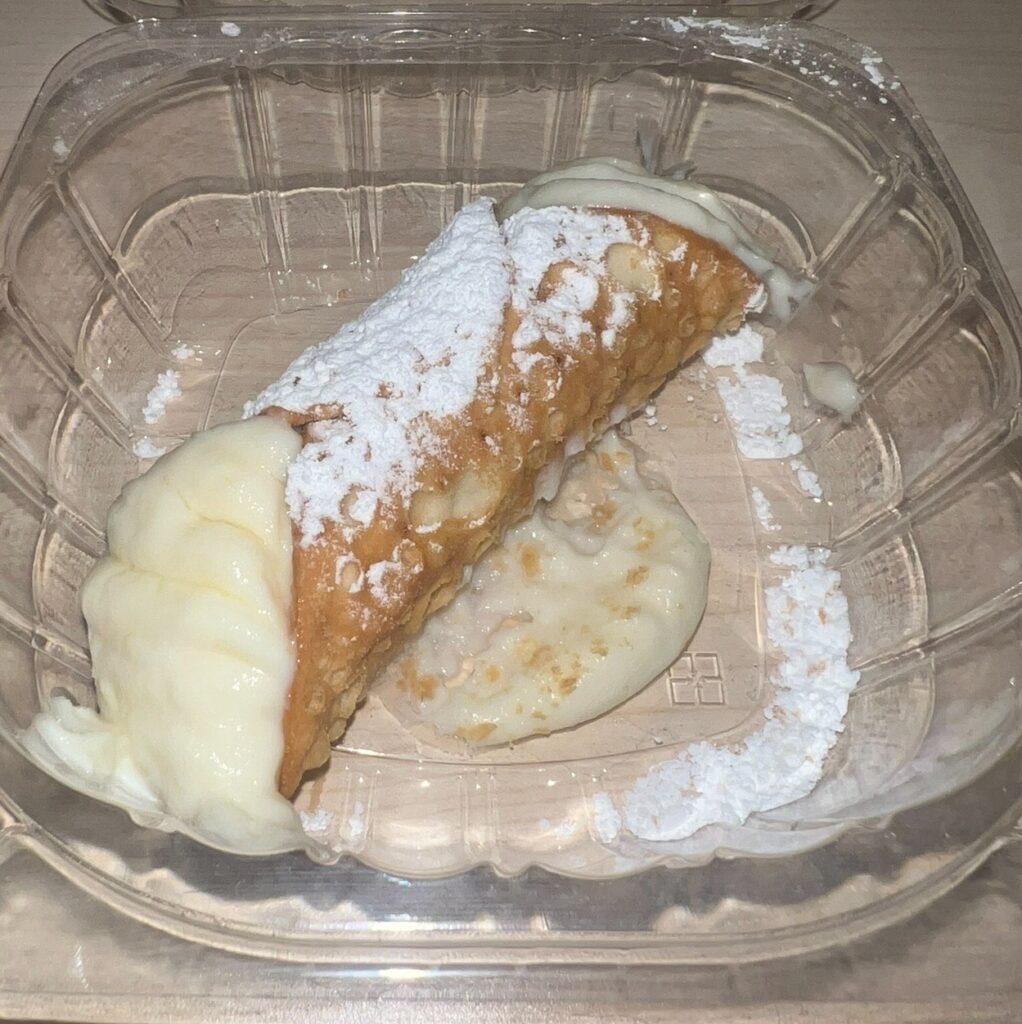 Classic cannoli filled with creamy ricotta cheese and dusted with powdered sugar, resting in a clear plastic takeout container.