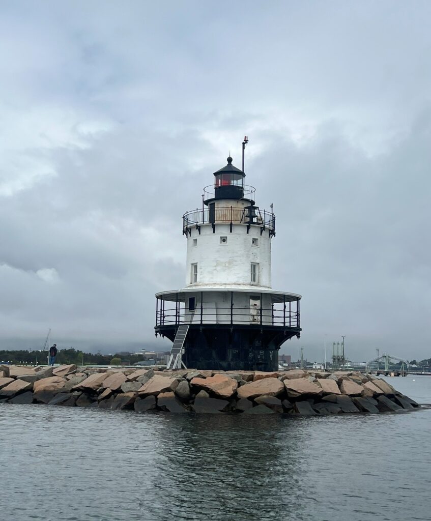 A solitary white and black lighthouse sits on a rocky breakwater surrounded by calm water under a cloudy sky. The cylindrical structure features a black railing and lantern room, with a ladder leading up from the rocks. A distant figure is visible on the breakwater, and industrial buildings can be seen faintly in the background across the water.