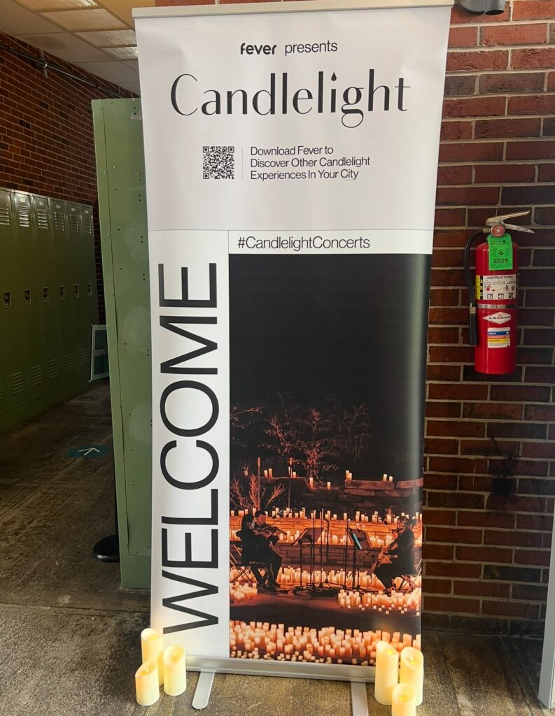 Vertical welcome banner for “Candlelight” concerts presented by Fever, positioned in a hallway with lockers and a fire extinguisher nearby. The banner features a nighttime concert scene with musicians playing surrounded by a sea of glowing candles, with artificial candles placed at its base for effect.