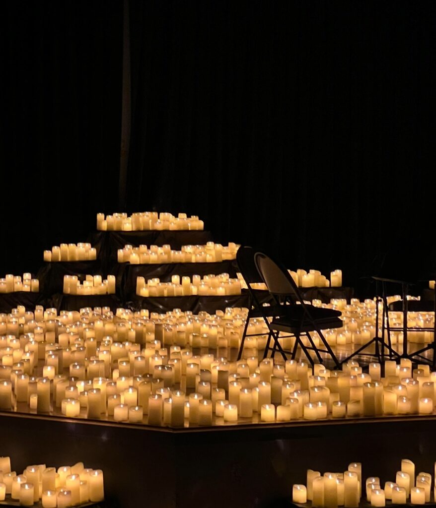 A dimly lit stage covered in hundreds of glowing flameless candles arranged in rows, including on tiered steps in the background. Two empty chairs and a music stand are positioned at the center, creating a warm, intimate ambiance for a live performance.
