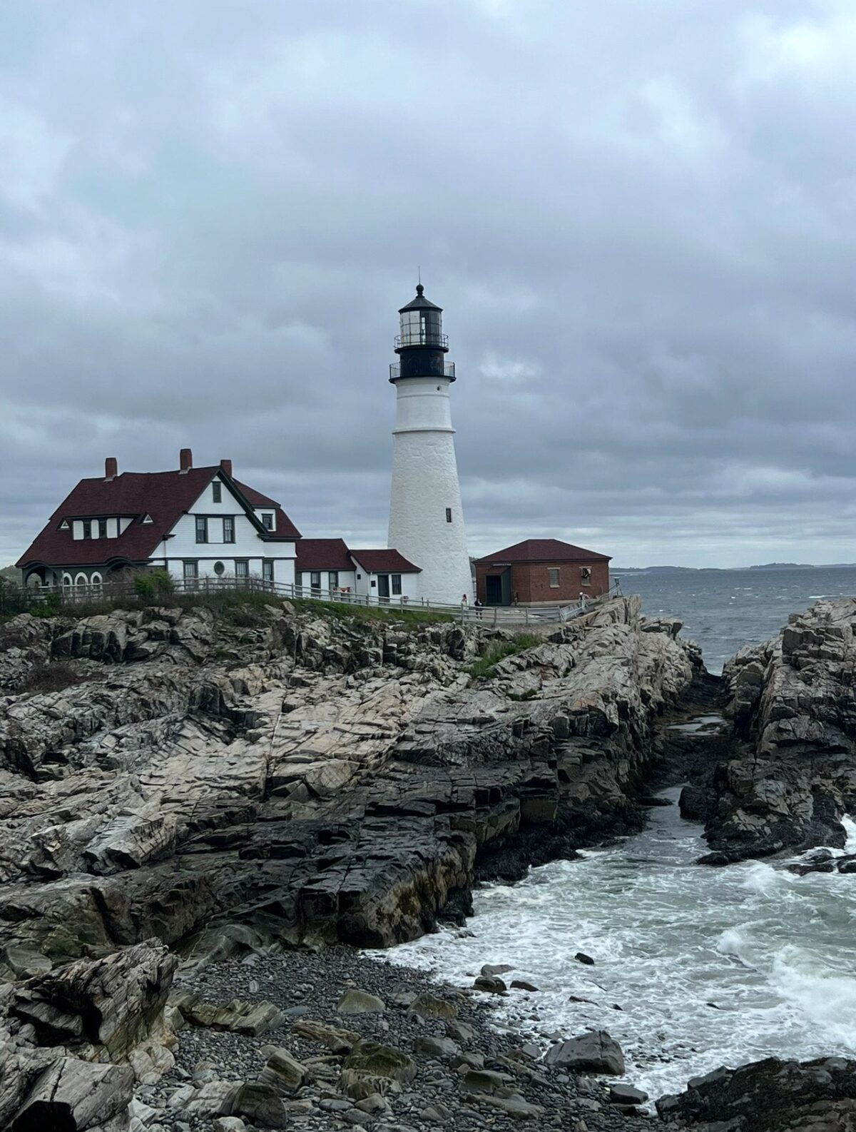 A scenic view of Portland Head Light, a historic white lighthouse standing on a rugged rocky coastline in Cape Elizabeth, Maine. The lighthouse is surrounded by red-roofed buildings and overlooks a choppy ocean under a cloudy sky.