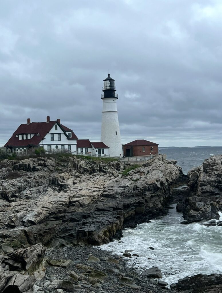 A scenic view of Portland Head Light, a historic white lighthouse standing on a rugged rocky coastline in Cape Elizabeth, Maine. The lighthouse is surrounded by red-roofed buildings and overlooks a choppy ocean under a cloudy sky.