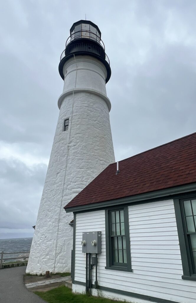 An upward view of the tall, white stone tower of Portland Head Light, showing its textured exterior and black lantern room. The corner of an adjacent white building with red roofing is also visible under a gray sky.
