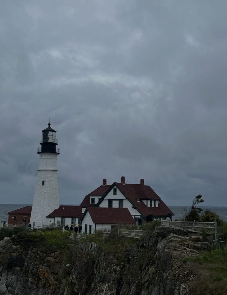 A moody coastal image of Portland Head Light with its adjacent white and red-roofed keeper’s house perched on a grassy cliff, set against a dark, overcast sky and the Atlantic Ocean in the background.