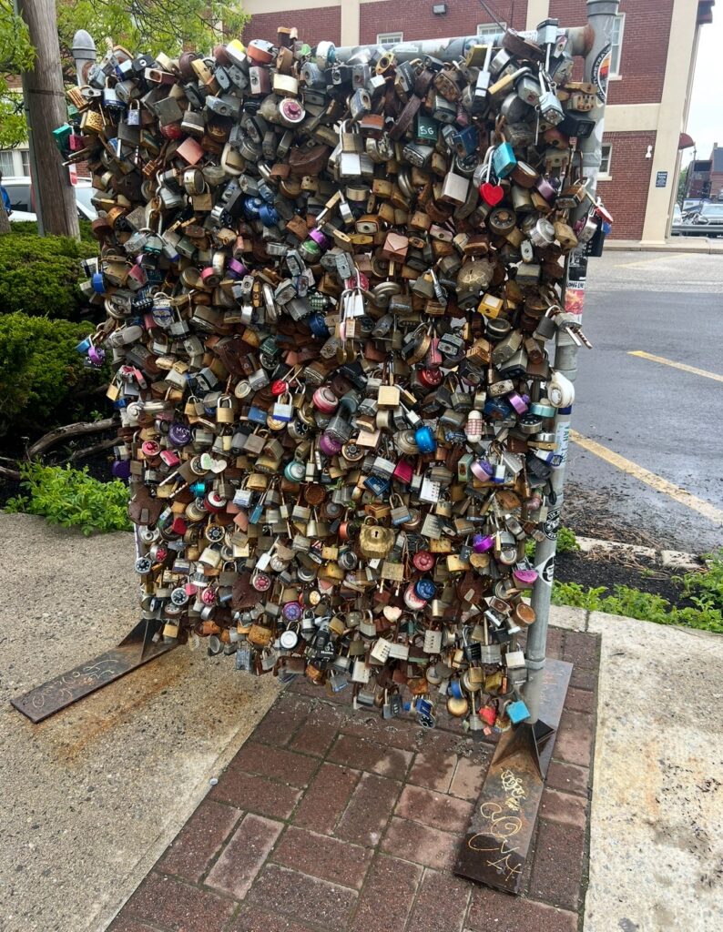 A dense metal structure filled with hundreds of padlocks, symbolizing love and commitment. The locks vary in size, color, and type—many are rusted, while some are decorated or engraved. The display is located on a sidewalk in an urban area near red brick buildings.