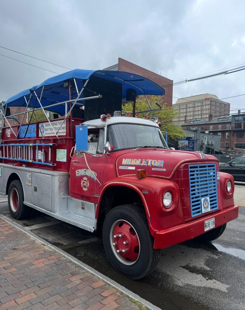 A bright red vintage fire truck labeled “Middleton Engine 3” is parked on a city street. The truck has been converted for sightseeing tours, with an open-air seating area covered by a blue canopy. The truck features chrome details and signage directing visitors to buy tickets online.