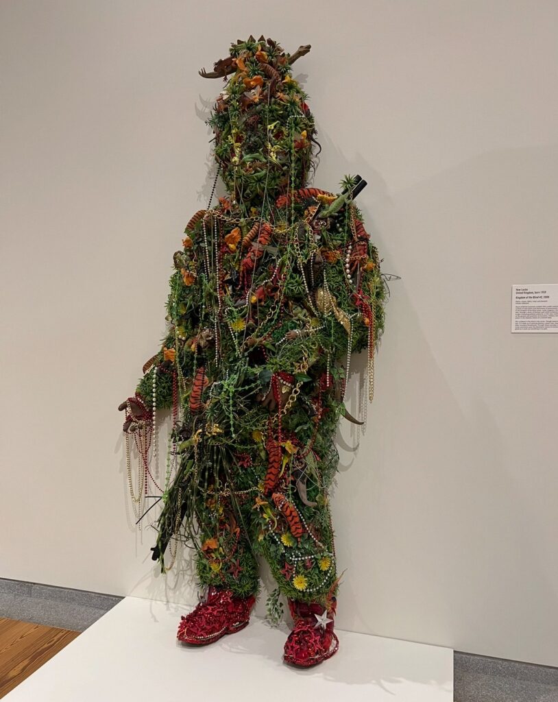 Lifelike human-shaped sculpture entirely covered in artificial plants, flowers, beads, chains, and toy animals. The figure wears red sequined shoes and stands against a white wall, creating a visually dense and whimsical blend of nature, fashion, and fantasy.