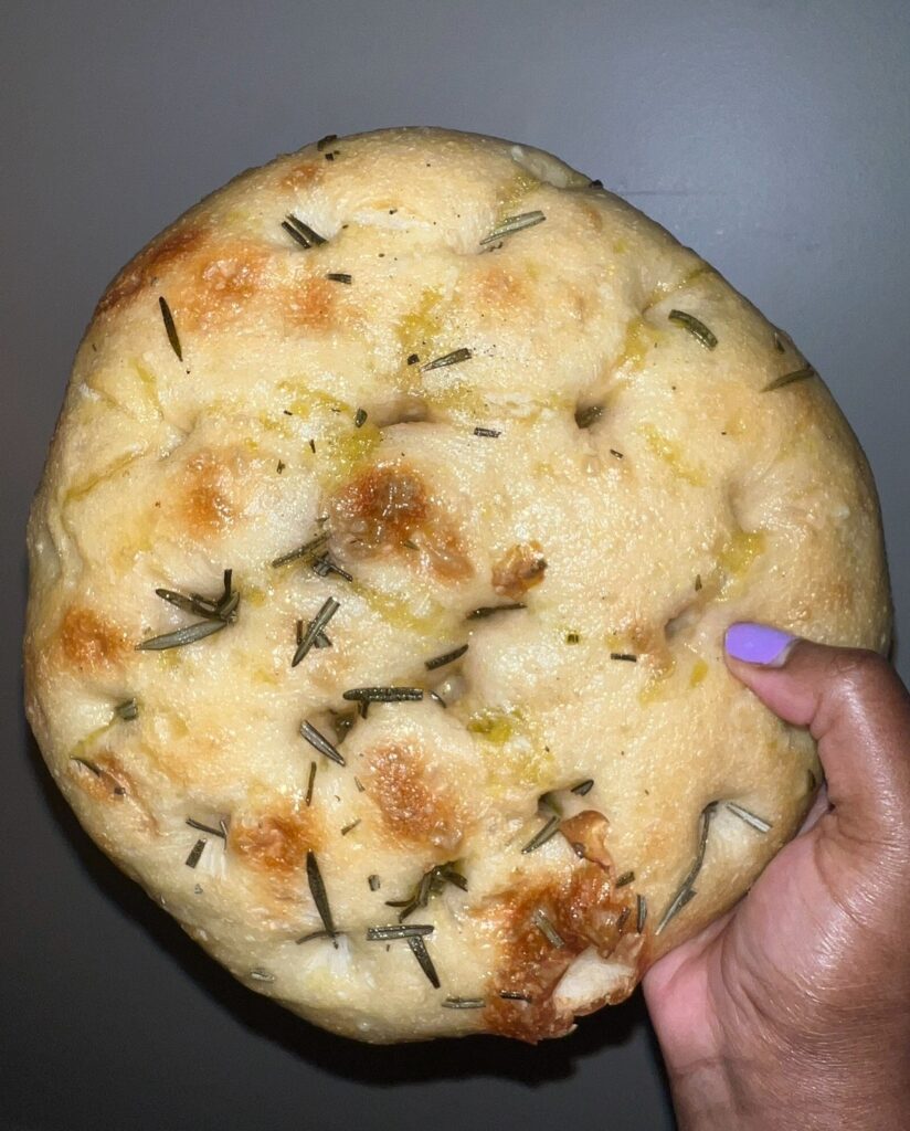 A hand holding a golden-brown round focaccia bread, topped with rosemary and olive oil, against a plain background.