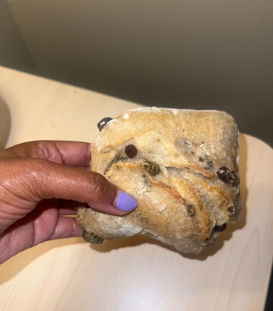 Close-up of a hand holding a rustic marinated olive bread roll with a golden crust and dusted with flour.