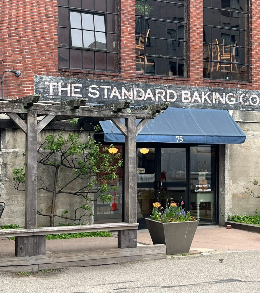 Front entrance of The Standard Baking Co. with a blue awning, rustic wood pergola, and colorful spring flowers in a planter outside a red brick building.