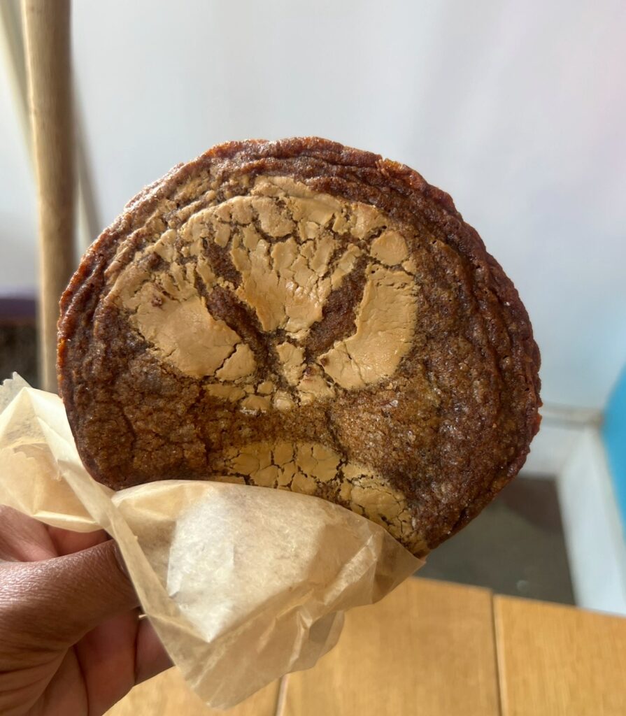 A hand holding a large, round, crinkly cookie with a caramelized edge and a unique crackled top that resembles an expressive face with furrowed brows. The cookie is partially wrapped in parchment paper.