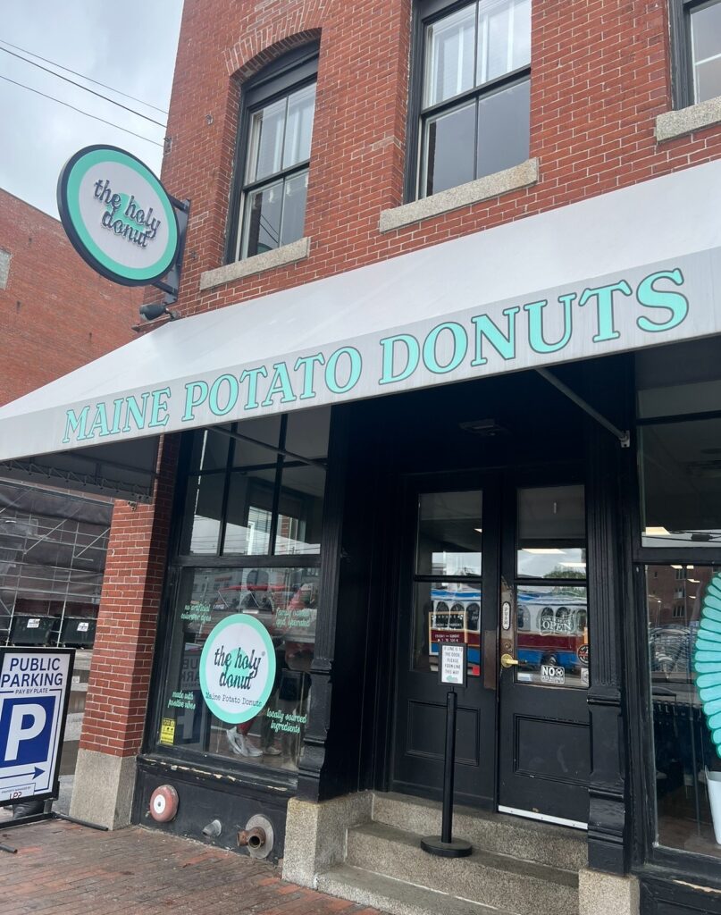 A street-level view of The Holy Donut storefront in Portland, Maine. The shop has a red brick exterior with large black doors and a white awning that reads “Maine Potato Donuts.” A round teal and white sign with the shop’s name hangs above the entrance.