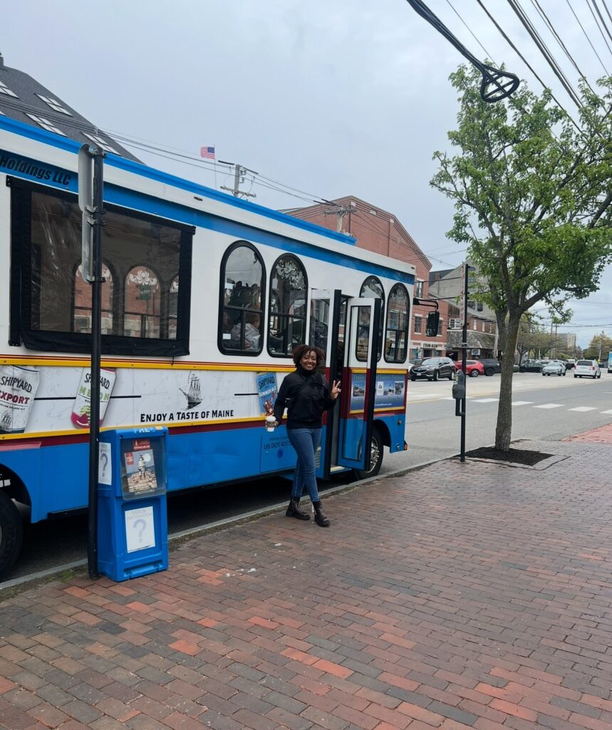 African American woman standing outside of a parked colorful trolley-style tour bus.