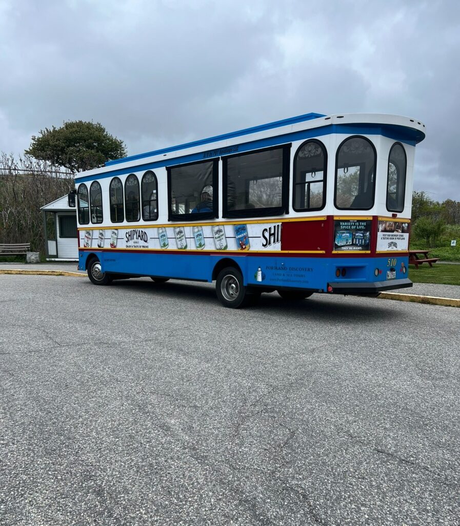 A colorful trolley-style tour bus parked on a paved road under a cloudy sky. The bus is branded with Shipyard Brewing Company advertisements and Portland Discovery land & Sea Tours. The bus is blue, white, and maroon with large windows, and several cans of Shipyard beer are pictured on the side. Trees, a bench and a small white structure are visible in the background.