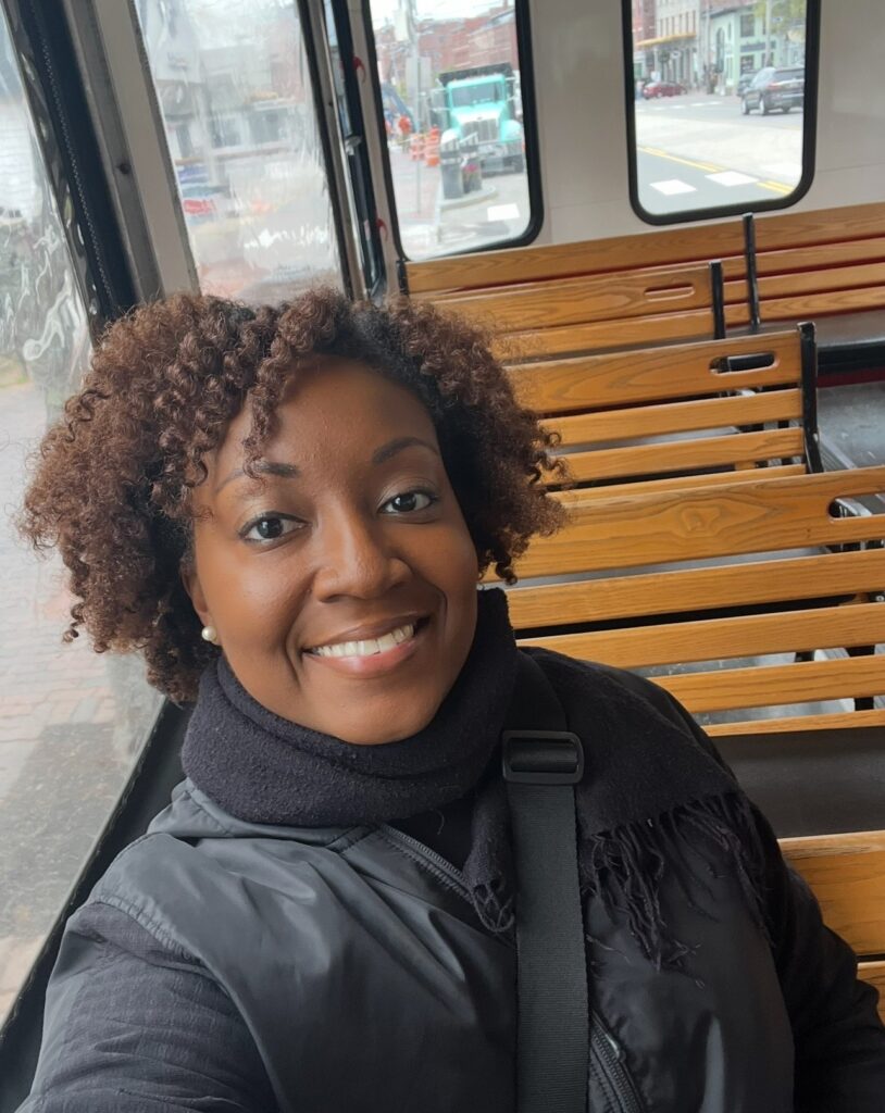 An African American woman with curly hair and a warm smile takes a selfie while seated on a wooden bench inside a trolley. She is wearing a black scarf and jacket, and the background shows the interior of the trolley with large windows and more wooden bench seating. Outside the trolley, a street scene with buildings and a teal truck is partially visible.