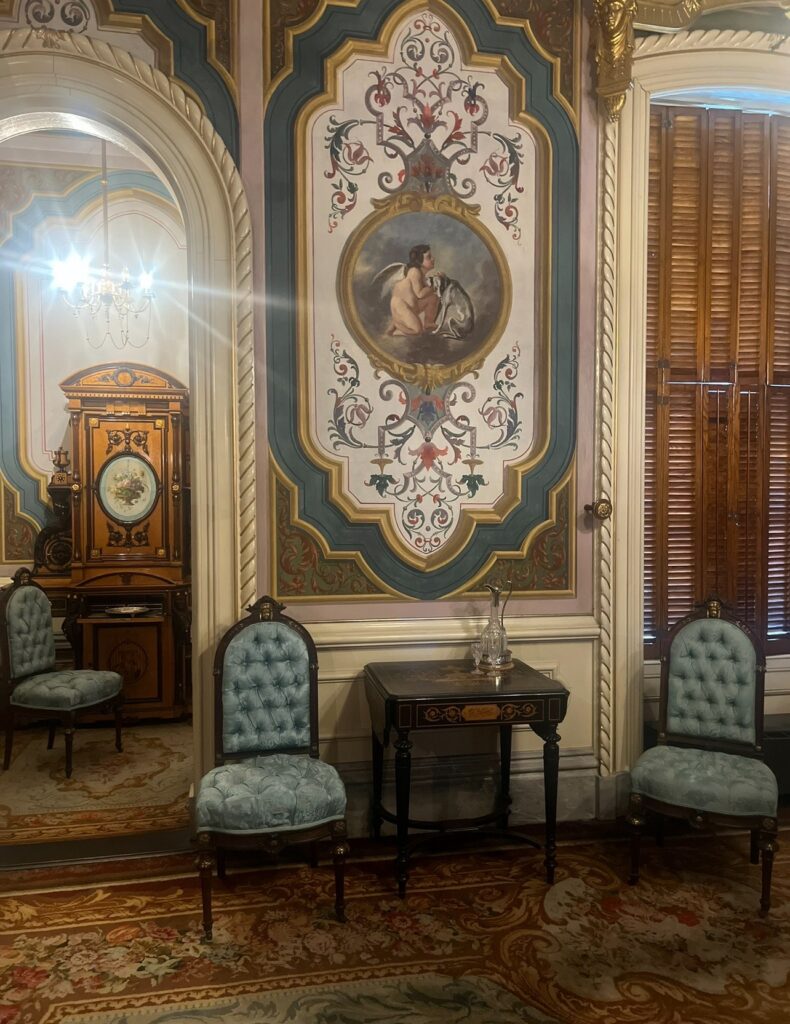A richly decorated Victorian-era room in Victoria Mansion with ornate wall paneling, angelic mural artwork, tufted blue chairs, a small decorative table, and wood blinds. A chandelier and antique clock are partially visible through the mirrored wall