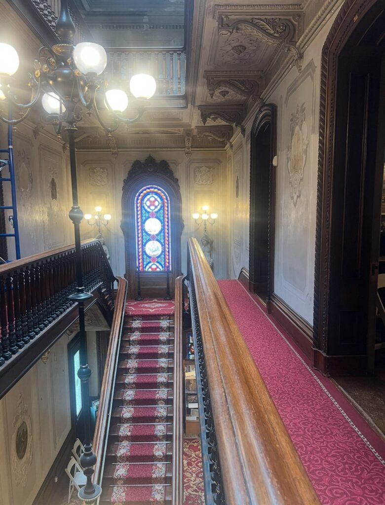 A grand staircase inside Victoria Mansion with red-patterned carpet, wooden banisters, and intricate ceiling molding. A large stained glass window illuminates the stairwell, flanked by ornate wall sconces and a chandelier.