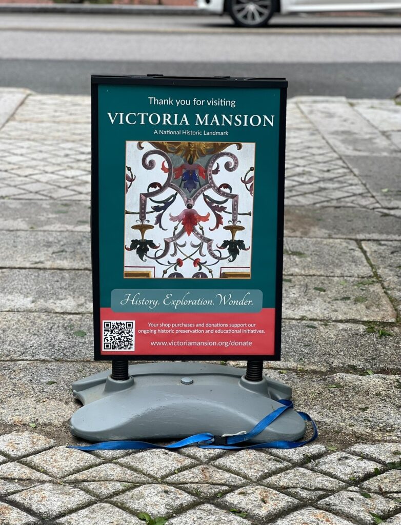 A standing sign thanking visitors for coming to Victoria Mansion, a National Historic Landmark. The sign features an ornate interior detail, a QR code, and a message promoting donations for preservation, placed on a cobblestone sidewalk near a street.