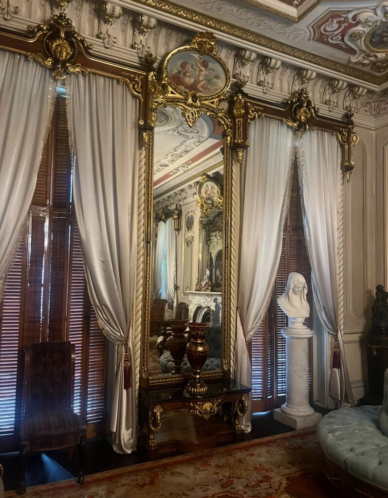 An opulent parlor scene in Victoria Mansion showcasing a large, gold-framed mirror with a painted cherub detail above it. The mirror is flanked by tall, pleated curtains, marble busts, and elegant furnishings, reflecting a lavish interior.