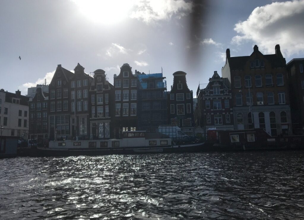 Canal houses at sunset in Amsterdam – A row of tall, narrow canal houses with traditional stepped gables face the water in Amsterdam. The sun is low in the sky, casting the buildings in silhouette, with shimmering reflections on the canal.