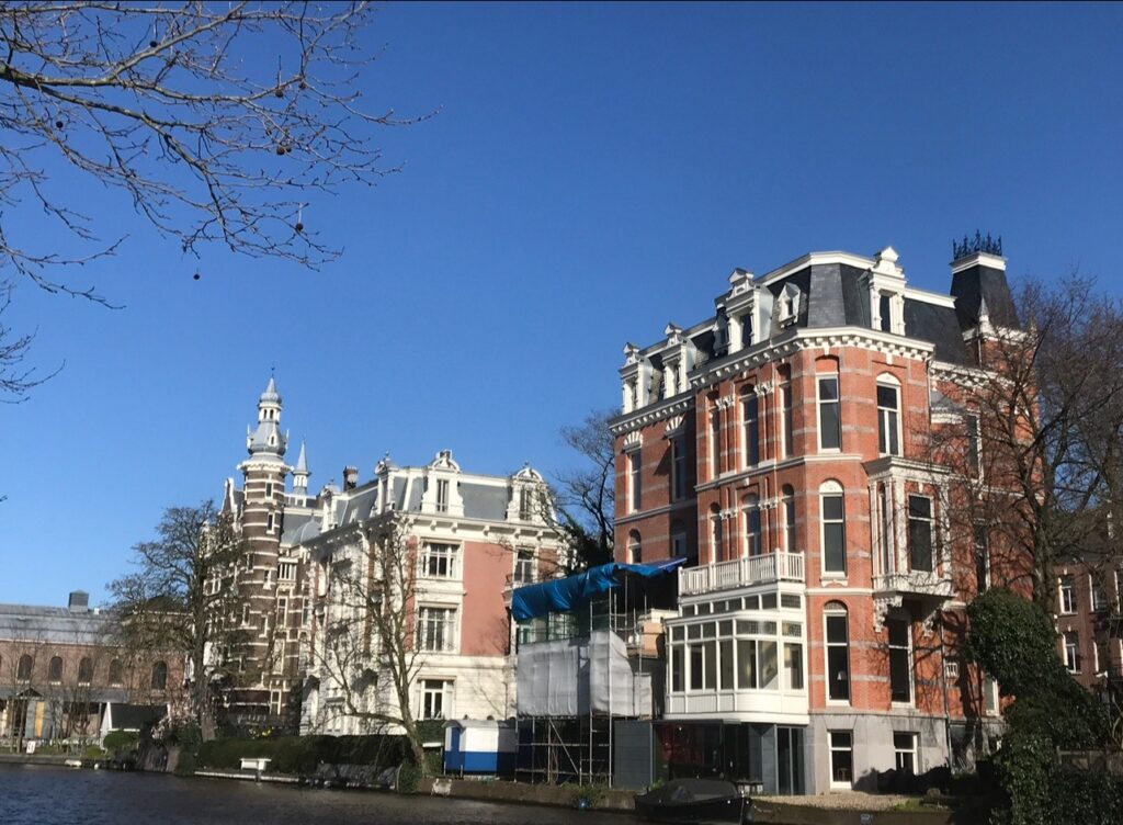 Historic canal houses in Amsterdam – Elegant brick mansions with ornate rooftops sit along a canal in Amsterdam. One building is wrapped in scaffolding, while the others showcase decorative gables under a bright blue sky.