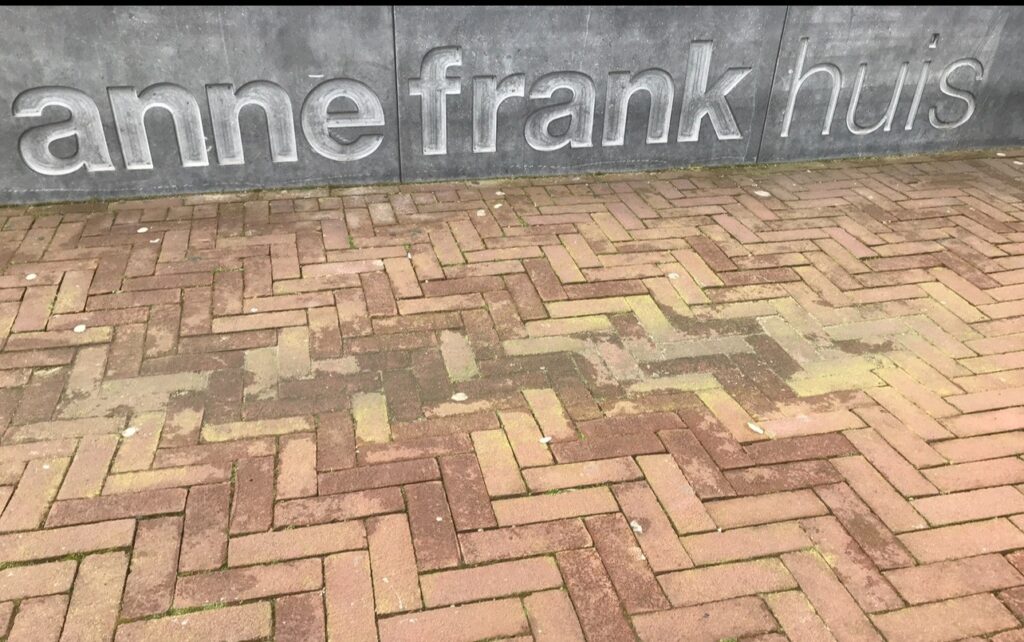 Anne Frank House sign – A close-up of the stone wall outside the Anne Frank House in Amsterdam, with the words “Anne Frank huis” engraved in large gray letters above a brick-paved walkway.