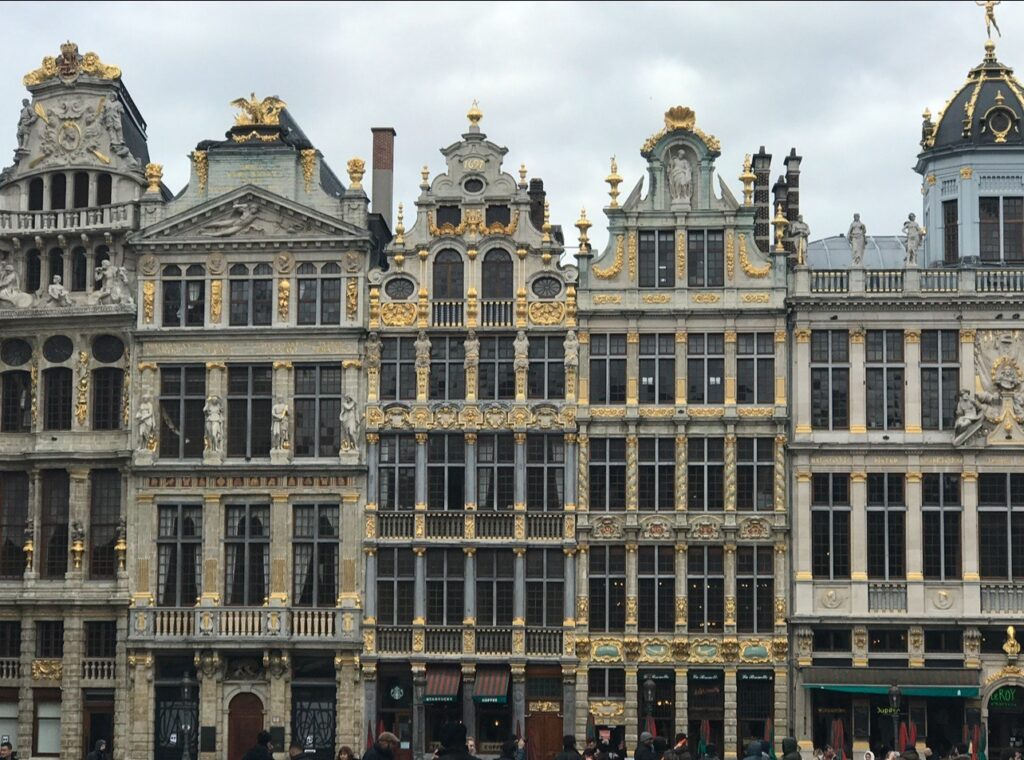 Grand Place guildhalls in Brussels – Ornate guildhalls in Brussels’ Grand Place with intricate facades decorated in gold detailing and statues. The richly adorned row of historic buildings stands under a gray sky.