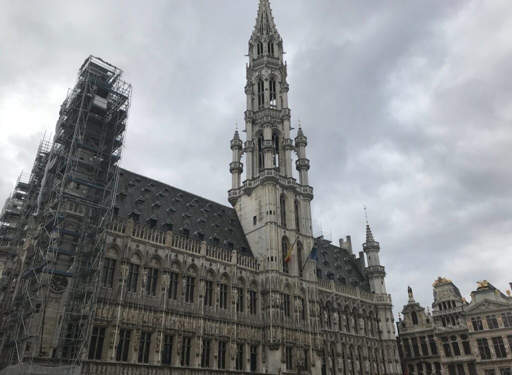 Brussels Town Hall – The ornate Gothic-style Brussels Town Hall, with its tall spire and detailed stone carvings, dominates the Grand Place. Part of the building is covered in scaffolding under a cloudy sky.