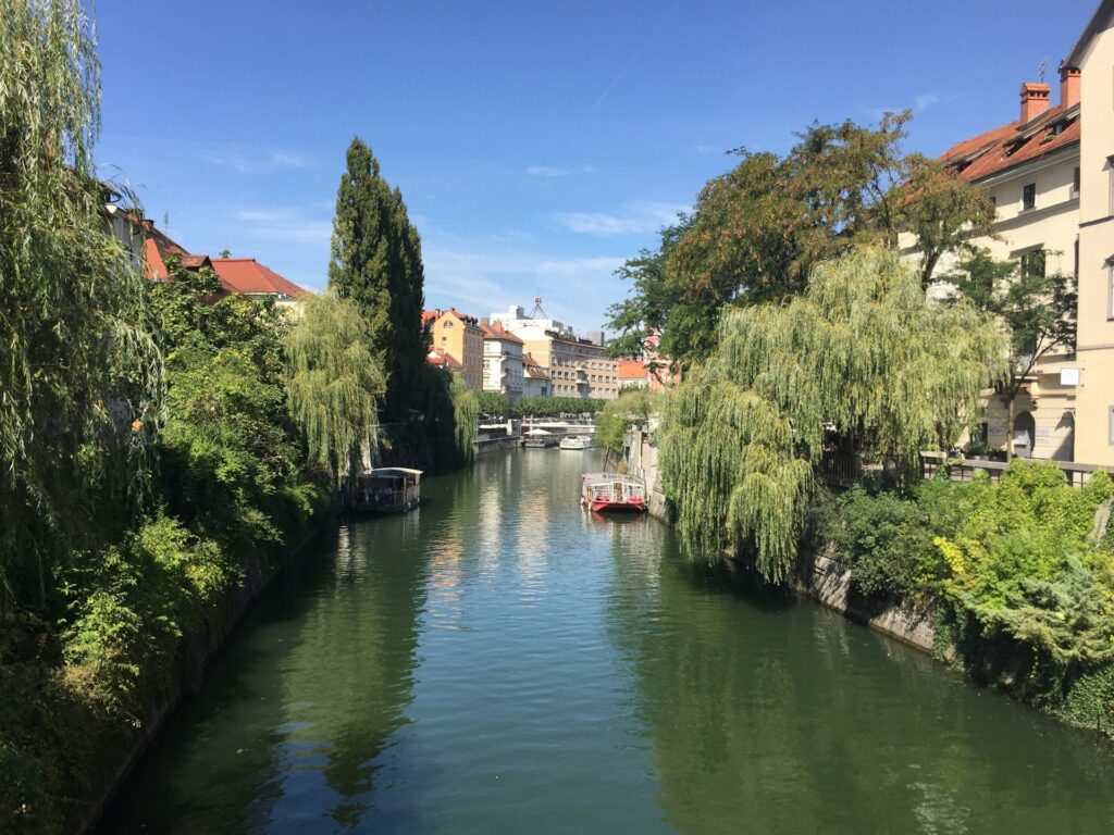 Scenic canal view in Ljubljana, Slovenia, lined with lush green willow trees and historic buildings with red rooftops under a clear blue sky, with small boats docked along the water.