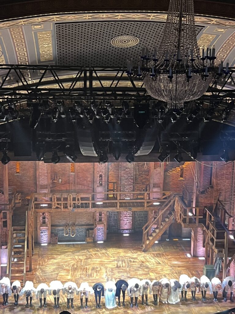 Cast of Broadway show taking a bow on stage beneath ornate theater ceilings, chandeliers and theatrical lighting, with a detailed wooden set in the background. 