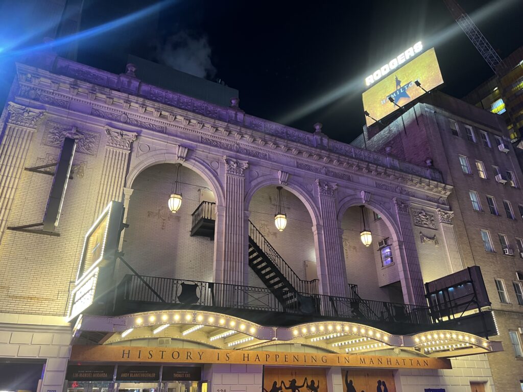 Exterior of the Richard Rodgers Theatre at night, illuminated with marquee lights and signage reading "History Is Happening in Manhattan," with Hamilton branding visible. 