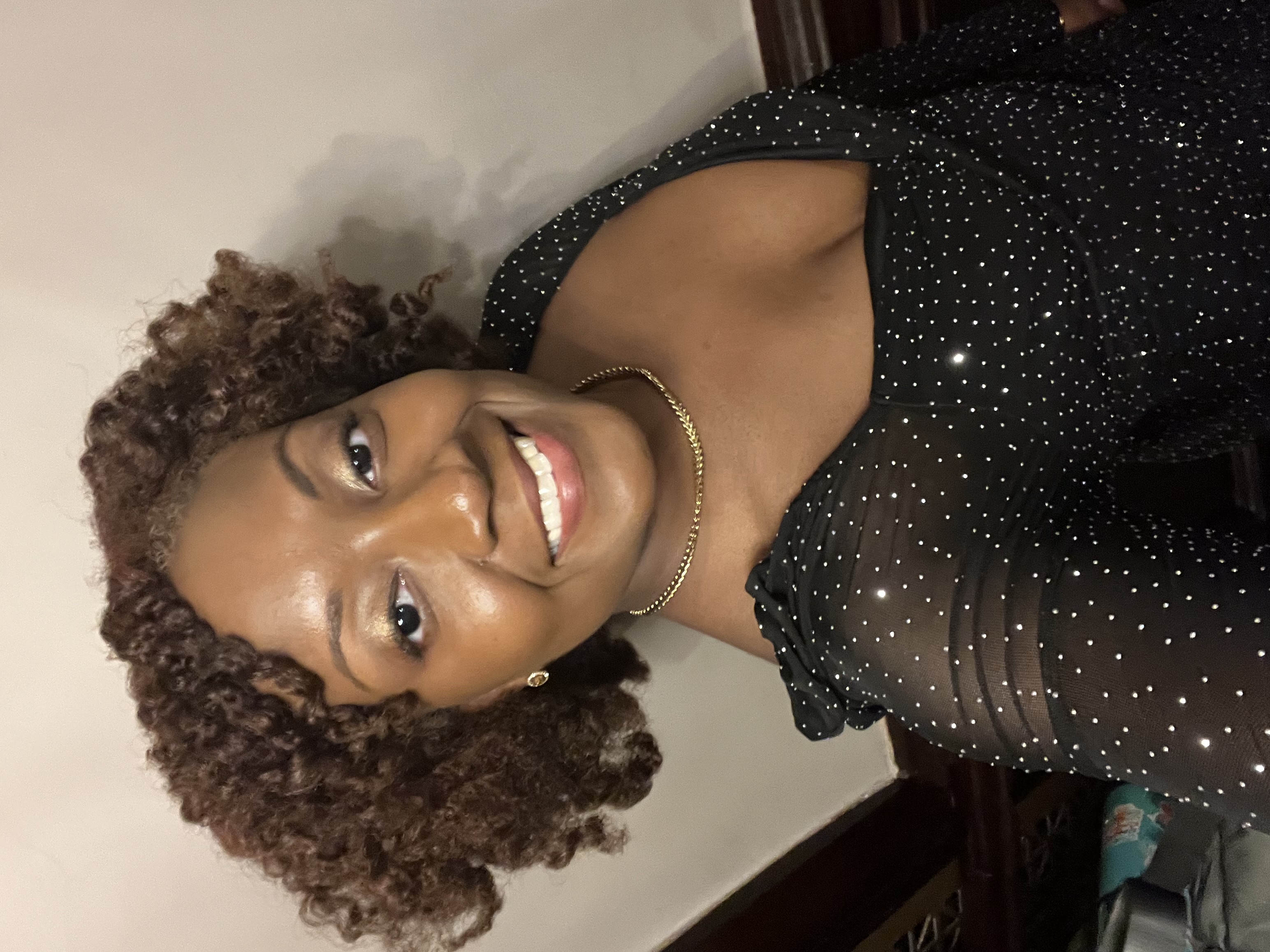 Smiling woman wearing a black sheer sparkle dress with a square neckline and gold jewelry, posing for a selfie indoors with softly curled hair and warm makeup. 