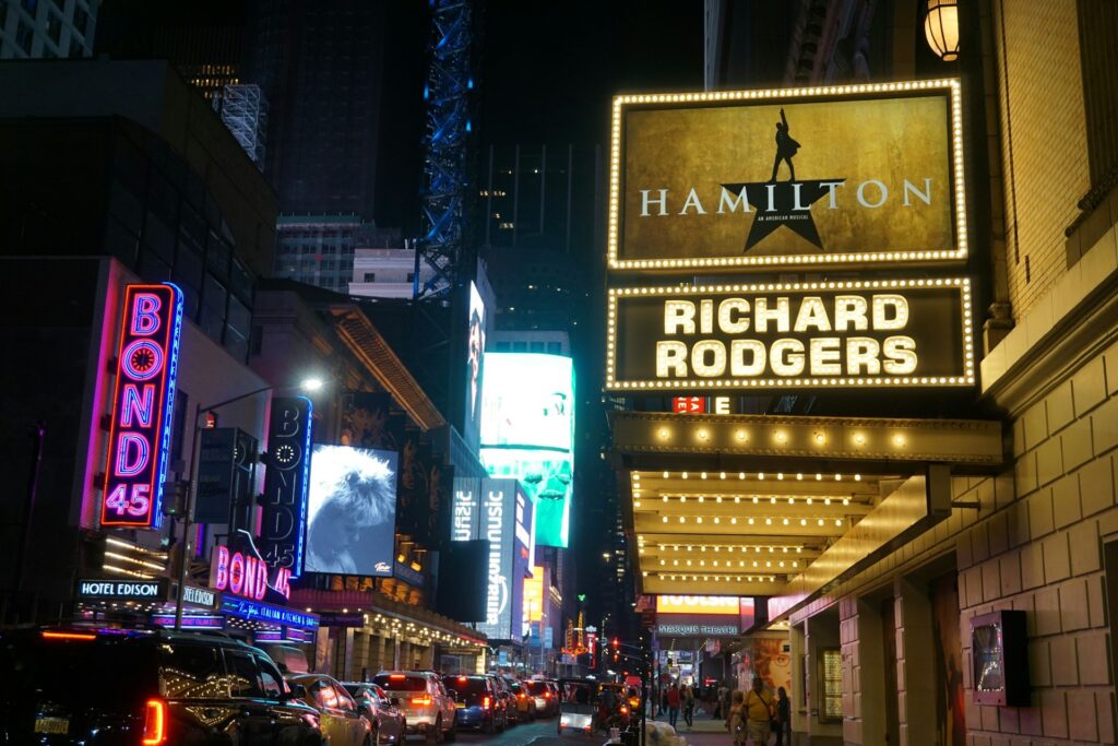 The Hamilton marquee glowing at night outside the Richard Rodgers Theatre on Broadway in New York City, with bright theater lights, city traffic, and surrounding Broadway signs.
